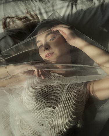 Portrait of Vika lying on a bed, partially covered with sheer fabric and lit by soft window light.