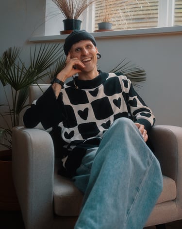 Cody on a grey chair wearing a beanie, baggy jumper, and baggy jeans, with plants in the background.