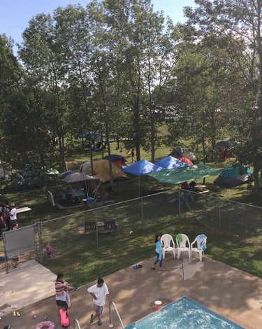 Crowded summer campground with tents under green trees next to a swimming pool and patio area.