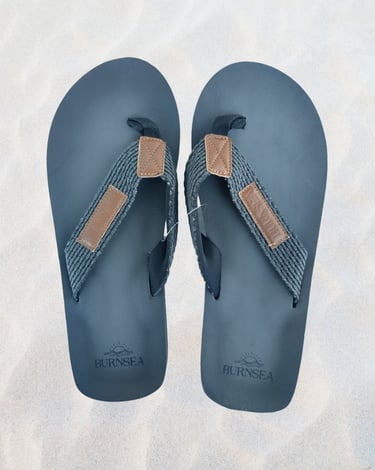 A pair of black Burnsea flip flops with grey fabric straps on a white sand background