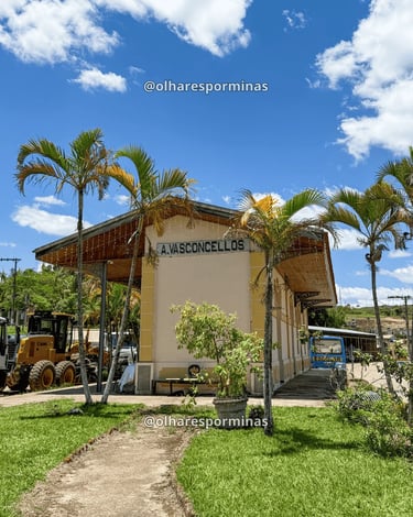 Fachada da estação ferroviária de Alfredo Vasconcelos sob céu azul com nuvens e palmeiras ao lado