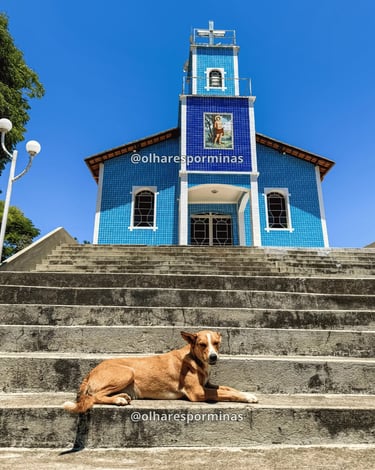 Igreja azul de Hermílio Alves MG com escadaria, cachorro e céu aberto
