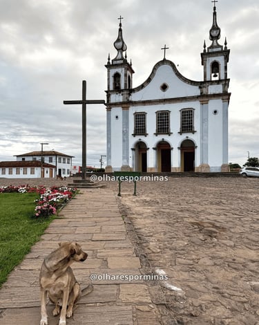Igreja histórica em Catas Altas MG com adro de pedra, cruz de madeira e cachorro em primeiro plano