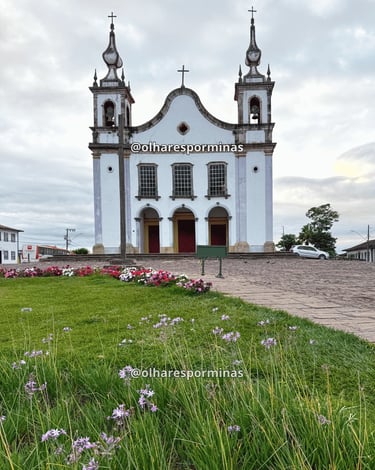 Igreja histórica em Catas Altas MG com jardim florido, torres coloniais e céu nublado ao fundo