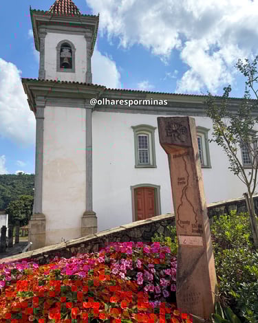 Toten da Estrada Real ao lado de um jardim florido em frente a uma igreja histórica em Santa Bárbara