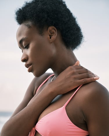 Side-profile portrait of Lauris wearing a pink swimsuit, photographed by the sea in natural light.