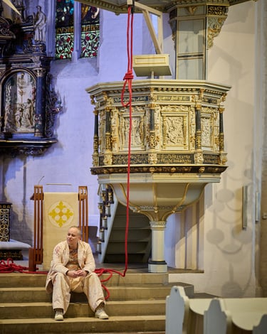 Ein Schauspieler sitzt in einer Kirche auf einer Treppe unter der Kanzel. Über ihm ein Galgenknoten.