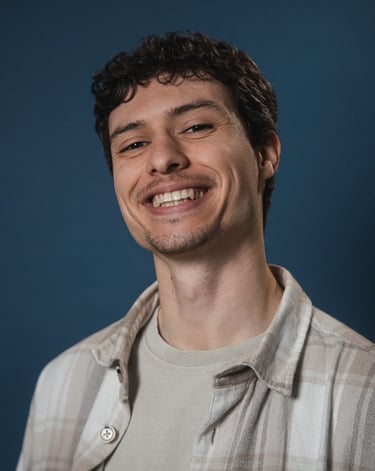 A smiling young man with curly hair and a plaid shirt posing against a blue background.