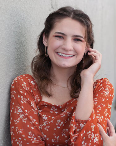 Brown haired woman in an orange dress with white flowers, posing in front of a white wall