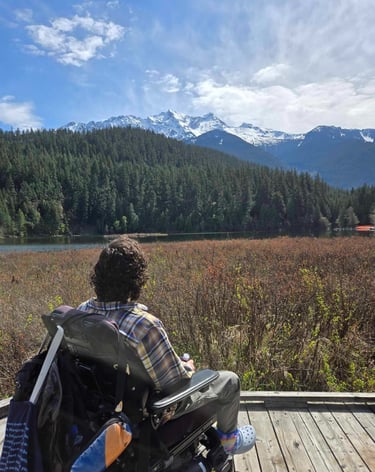Sean in his power wheelchair on a lakeside boardwalk looking at Mt Currie in BC