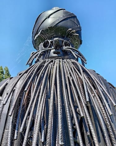 Black metal garden sculpture with a rebar beard and spider webs against a blue sky.