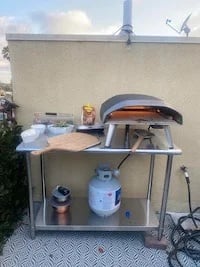 A pizza oven and prep tools sitting on a stainless steel table with a gas tank stored on the bottom shelf outdoors.