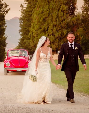 Sposi che camminano per mano lungo un viale alberato, sorridenti davanti alla loro auto d’epoca
