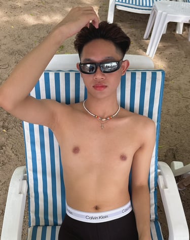 Hnadsome young man wearing black underwear and sitting on a beach chair with right hand on his head and wearing sunglasses