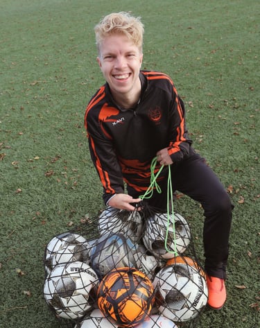 Smiling soccer coach on a turf field holding a mesh ball bag full of footballs for sports training.