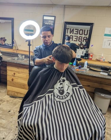 A student barber giving a haircut with electric clippers in a barber college setting.