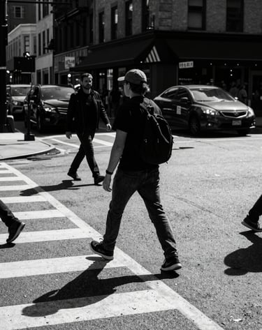 A dynamic street photography shot in a North American urban setting, captured in high-contrast black and white, showing a busy intersection with dramatic shadows and deep charcoal tones.