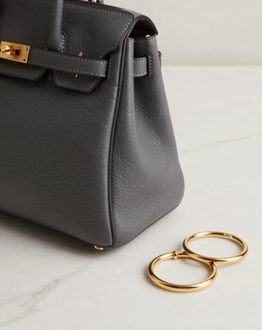 Close-up detail of a deep slate grey designer handbag and soft gold jewelry placed on a minimalist soft off-white marble surface. Elegant and sophisticated lighting in a North American / Western European design studio.