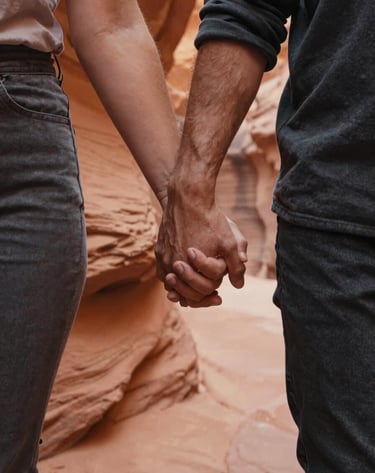 Close-up cinematic shot focusing on a couple's entwined hands as they walk through a North American / US canyon. The warm sun-drenched light catches the textures of the terracotta rock walls and their casual charcoal clothing. The mood is intimate and peaceful.