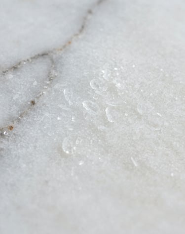 Extreme close-up macro photography showing the intricate crystalline texture of a white marble surface. Soft, diffused lighting highlights the translucent quality of the stone. Subtle gray veins meander across the surface. The aesthetic is clean, editorial, and sophisticated.