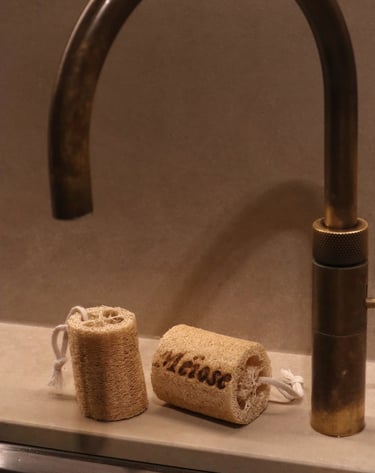 Two natural loofah sponges with cotton loops sit on a stone counter next to a vintage brass faucet.