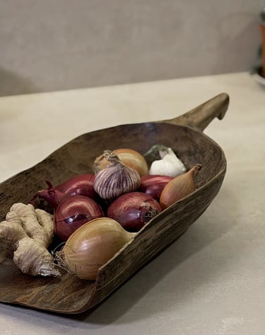 Rustic wooden scoop filled with red onions, shallots, garlic, and fresh ginger root on a stone countertop.
