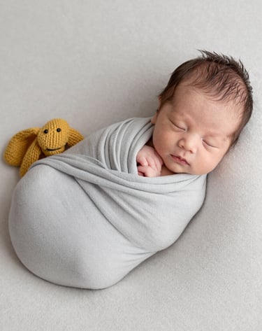 Minimalist newborn baby photography. A peaceful infant wrapped in a soft light grey blanket, resting on an off-white textured surface. Artistic, warm lighting with a subtle mustard yellow knitted accessory. Professional and inviting mood.