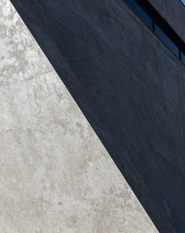 A detailed close-up shot of architectural textures. A smooth light silver grey concrete surface meets a dark charcoal grey metallic finish. Harsh sunlight creates a dramatic diagonal shadow across the frame, emphasizing the high-contrast aesthetic of a modern South American / Brazilian building.