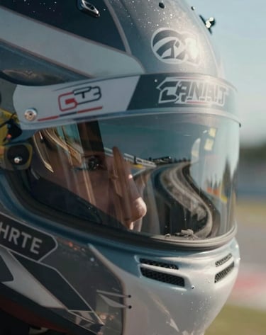 A macro shot focusing on the visor of a racing driver's helmet. The dark gray visor reflects the distorted curve of a famous North American / US racetrack under a clear sky. Intimate and intense focus.