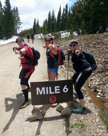 Three Spartan Race athletes posing by a Mile 6 sign on a mountain hiking trail with pine trees.