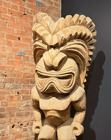 Hand-carved wooden Tiki statue with intricate facial details against a red brick wall.