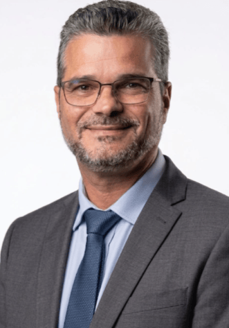 Professional headshot of a smiling businessman in a grey suit and blue tie with glasses.