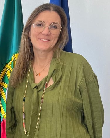 A smiling woman with glasses and a green blouse standing in front of the flag of Portugal.