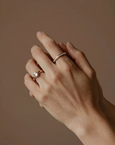 Close-up detail photography of two hands interlocked, wearing North American / US style wedding jewelry. The background is a soft focus of muted earth brown and terracotta tones. Warm, cinematic lighting.