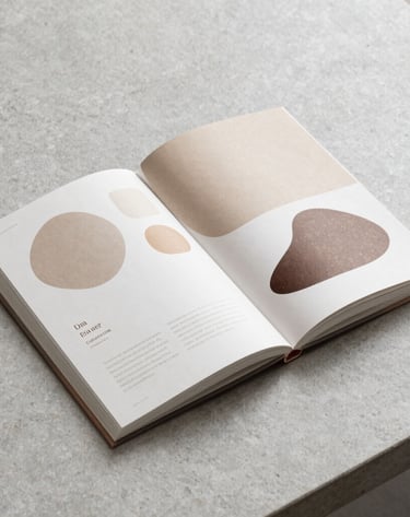 A minimalist brand guidelines book open on a light grey stone desk. The pages display organic shapes and an earthy color palette. Professional photography, clean and functional composition.