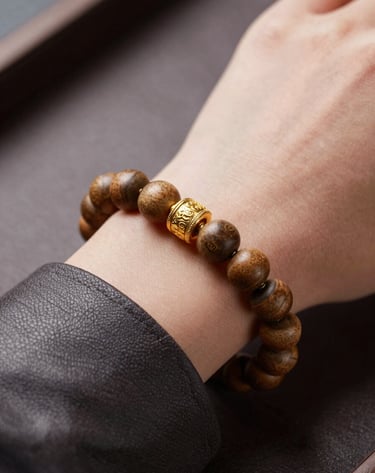 Close-up detail of high-end accessories: a mix of gold and traditional wooden beads on a wrist, resting on an expensive dark leather surface. Colors include #8D765D and #3D2C20. Shallow depth of field, very premium look.