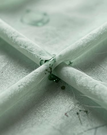 A macro photograph focusing on the intersection of ink and silk fiber. The ink bleeds slightly, creating a nebulous, atmospheric texture in muted sage green. The lighting is bright and airy, emphasizing the translucency of the material.