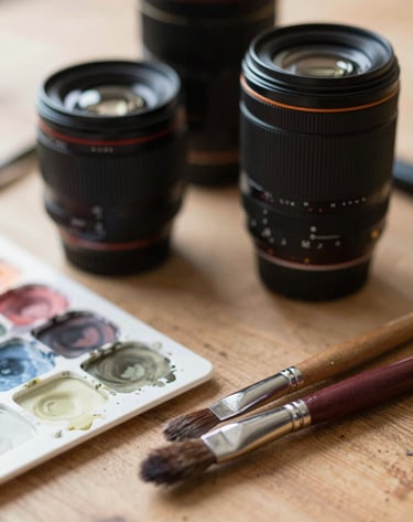 A close-up shot of professional camera lenses and watercolor brushes on a wooden surface, soft atmospheric lighting, highlighting the craftsmanship and tools of the artist, palette includes #6F5E52 and #F5F2EF.