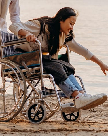 Donna in sedia a rotelle sulla spiaggia, accompagnata, mentre tocca l’acqua del mare