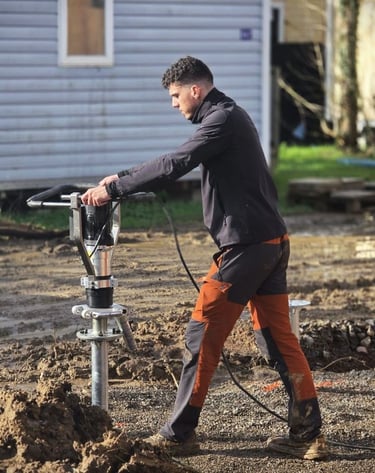 A construction worker using a power driver to install helical screw piles into muddy ground.