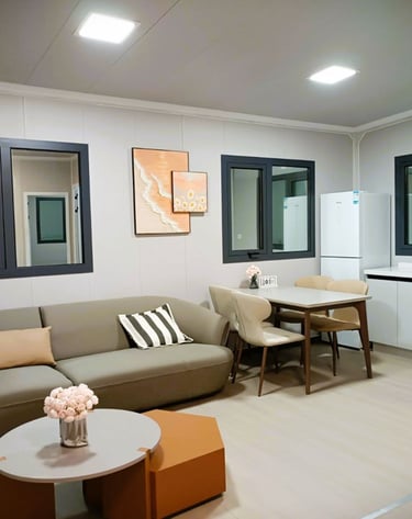 Modern studio apartment with a grey sofa, minimalist dining set, and white kitchenette with a refrigerator.