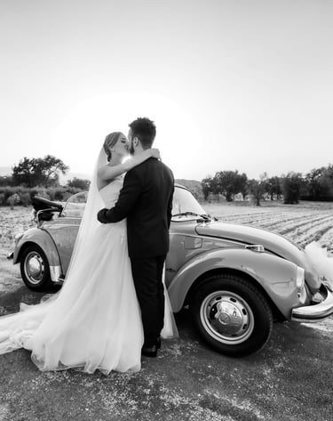 Sposi che si abbracciano accanto a un’auto d’epoca decorata, in un paesaggio di campagna al tramonto
