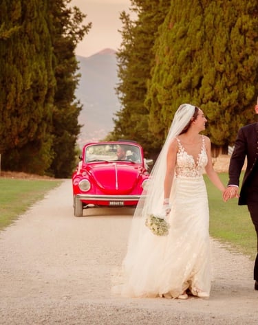 Sposi che camminano per mano lungo un viale alberato, sorridenti davanti alla loro auto d’epoca