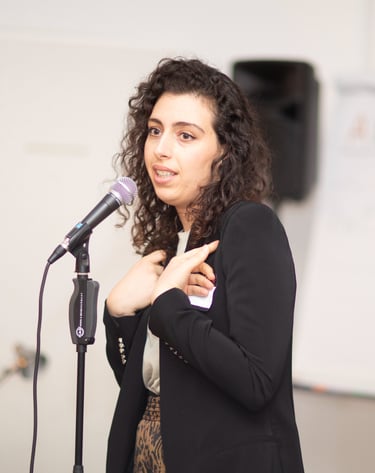 Woman behind microphone, making hand gestures, flipchart in the back, seminar setting.
