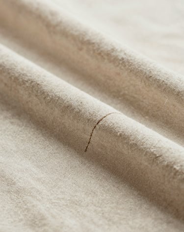 An intimate, close-up photograph of delicate hand-stitched detailing on luxury organic cotton fabric. The colors are muted warm beige and soft cream. The composition is extremely clean, minimalist, and focused on pure craft and material excellence, shot under natural daylight.
