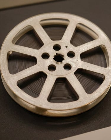 Close-up of a vintage 35mm film reel resting on a dark surface, light catching the metallic edges in muted gold tones, soft off-white ambient glow, evocative of a museum archive in Angola.