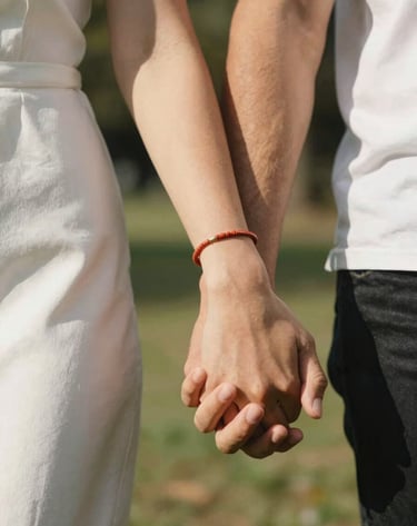 A detailed close-up shot of two people holding hands during a walk in a North American / US park. The lighting is sun-drenched and warm, highlighting terracotta jewelry and soft white fabric textures. Shallow depth of field.