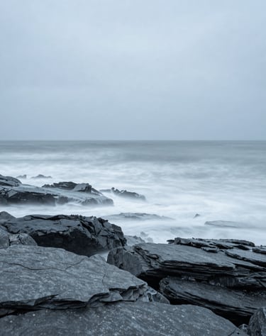 A minimalist long exposure shot of waves hitting a slate gray rocky shore. Soft misty texture, light blue gray sky, Global / General coastal scene.
