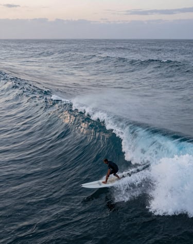 A high-speed drone shot capturing a surfer on a massive North American wave at dawn. Muted slate blue water tones and pale azure highlights on the spray.