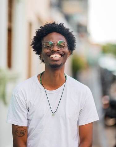 Homem preto e magro, de cabelo crespo. Usa óculos de armação fina e redonda. Tem o sorriso largo.